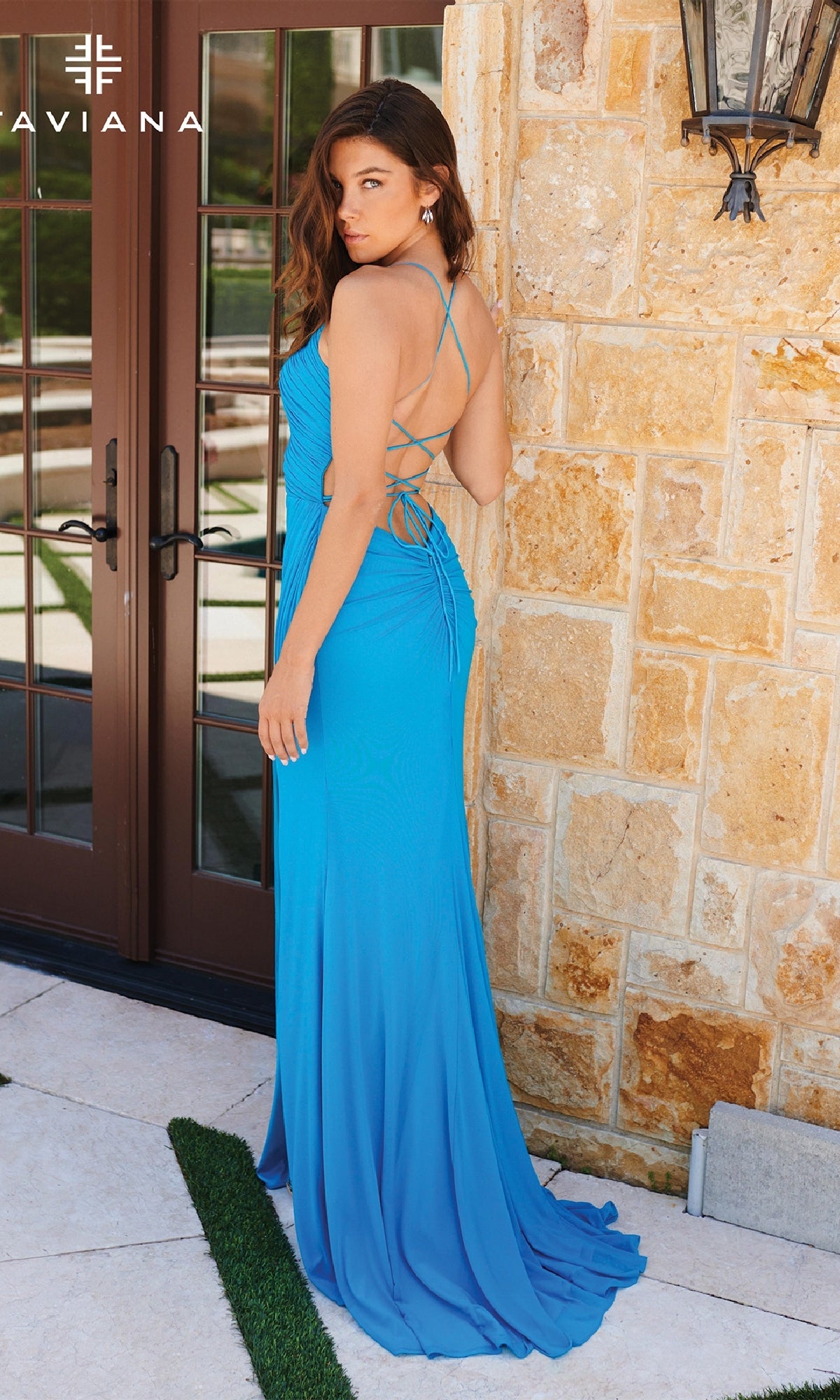 A woman stands near a stone wall and glass doors wearing the Faviana 11267, a bright blue, floor-length prom dress featuring an open back with crisscross straps and a lace-up detail.